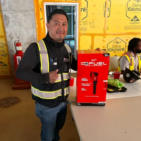 Construction worker in safety vest posing next to a Milwaukee M18 Fuel power tool box on a table inside a building under construction.