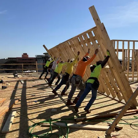 Construction workers lifting a wooden frame on a building site under clear blue sky.