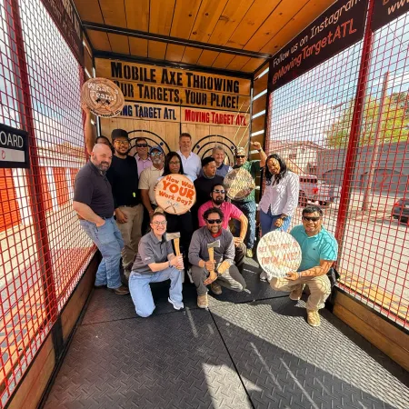 Group of people posing with axes and target signs inside a mobile axe throwing booth with wooden and metal walls.