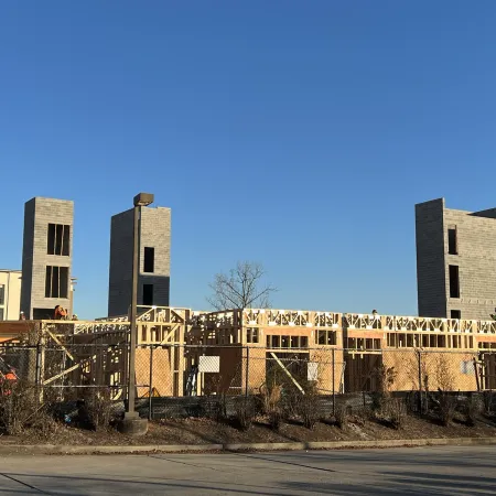 Construction site with wooden framing and concrete towers under clear blue sky, surrounded by fence and roadside signs.