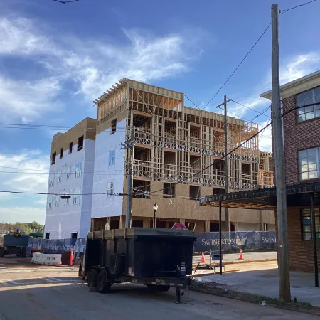 Mid-construction multi-story building with exposed framing and white wrap under a clear sky near a brick building.