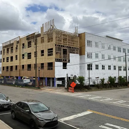 Large multi-story building under construction with wooden framing visible and parked cars on the street.