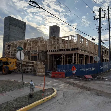 Wooden frame of a multi-story building under construction at a corner site with a crane and barriers around it.