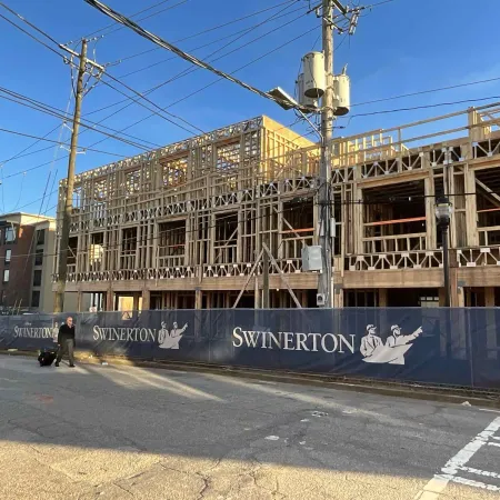 Multistory wooden frame building under construction behind Swinerton branded fence on sunny day.