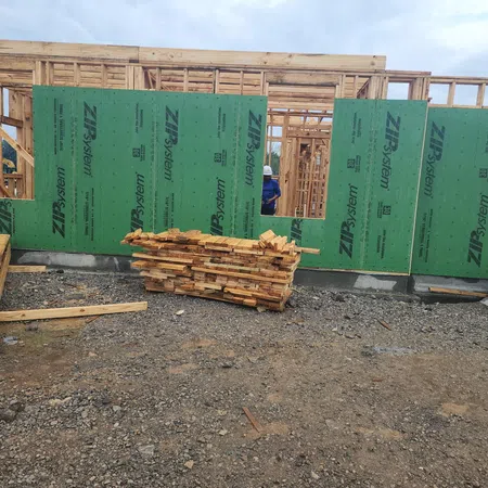 Partially constructed wooden house with green ZIP System sheathing panels and stacked lumber on muddy ground.