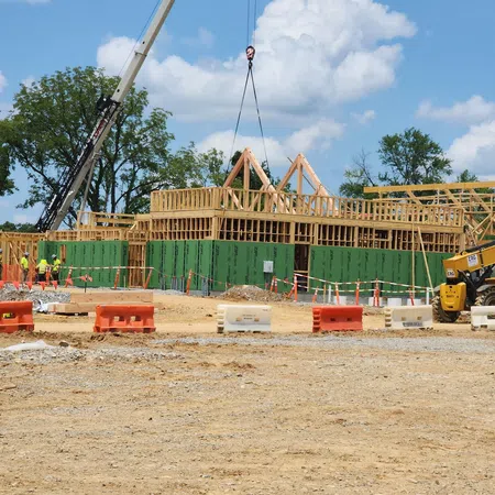 Construction site with wooden framing being lifted by crane and workers in safety vests on a sunny day.