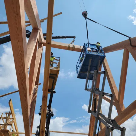 Construction workers in lifts assembling large wooden beams on a frame structure under blue sky with clouds