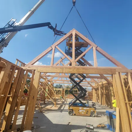 Wooden house frame construction with workers using a crane and lift under clear blue sky.