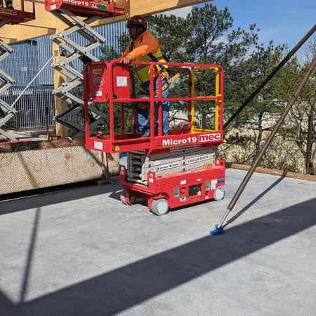 Construction worker in safety gear on a red scissor lift under wooden beams at an outdoor building site.