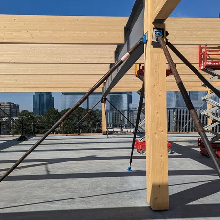 Wooden beam framework supported by metal braces at a construction site with city buildings in the background.
