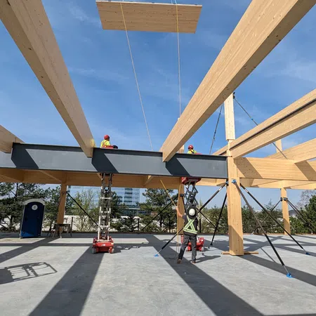 Construction workers lift wooden panel with crane inside timber frame structure under blue sky.