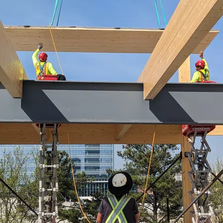 Construction workers guide a large wooden beam into place using harnesses and scissor lifts on a sunny day.