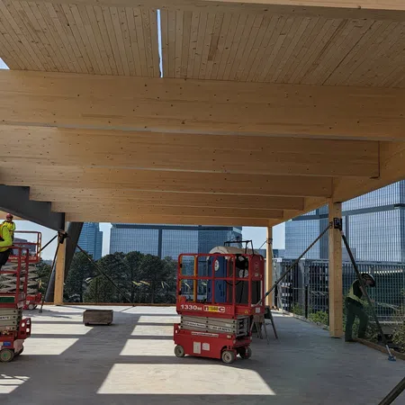Construction site with large wooden beams, workers, and equipment under a clear sky in an urban setting.