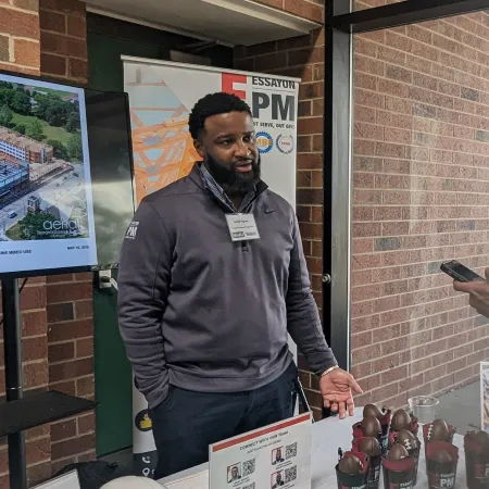 Two men at a trade show booth displaying promotional items and visuals in a brick-walled venue.