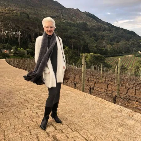 Elderly woman in white coat and scarf standing on vineyard path with mountains and cloudy sky in background