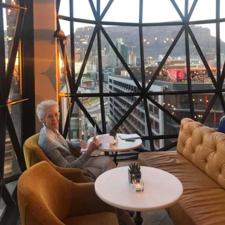 Elderly woman reading in a cozy lounge with panoramic city views through a geometric glass window.