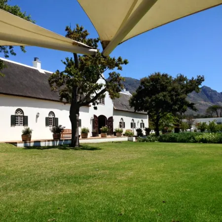 Traditional white farmhouse with thatched roof surrounded by green lawn and trees under a clear blue sky.