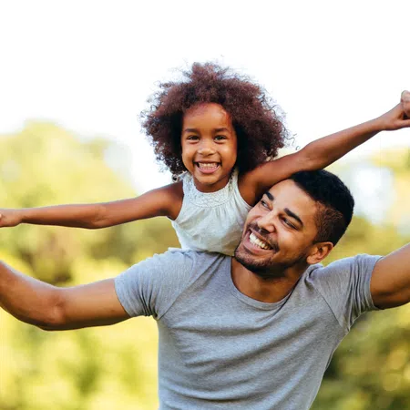 Happy father giving piggyback ride to smiling daughter outdoors with blurred green trees in background