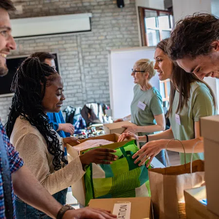 Volunteers smiling and packing donation boxes and bags in a bright community center setting.