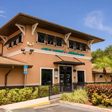 Exterior of Evora Women's & Children's Care clinic with palm trees and landscaped bushes under blue sky.