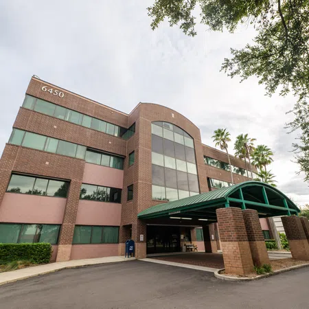 Four-story brick office building with green awning entrance, large windows, and surrounding trees under cloudy sky