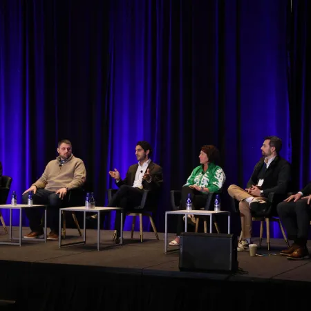 Six professionals seated on a stage panel discussion with blue curtains and small tables with water bottles.