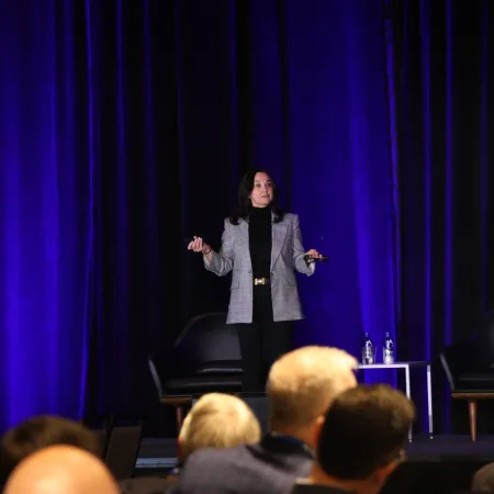 Woman in gray blazer presenting on stage with purple curtains and audience in foreground