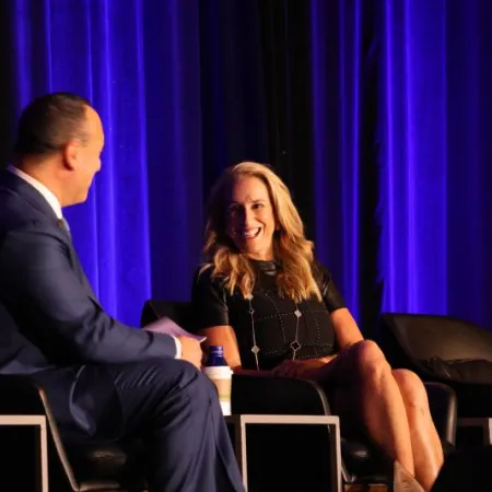 Man and woman in formal attire engaged in a lively conversation on stage with purple curtains in the background.