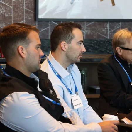Three men attentively listening at a professional conference wearing name tags and lanyards in an indoor setting.