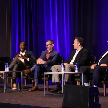 Five panelists engaged in discussion on stage with purple curtain backdrop at a professional conference event.
