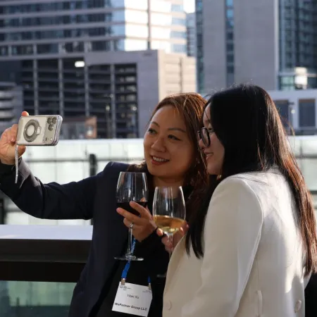 Two women in business attire taking a selfie on a rooftop with wine glasses and city buildings in background