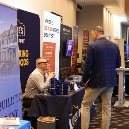Two men at a business expo booth with banners and promotional materials focused on design and construction.