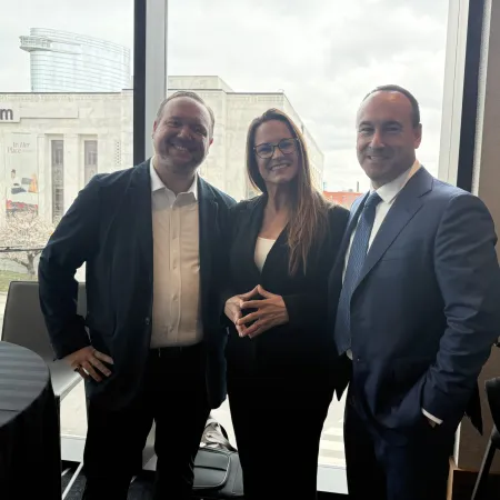 Three professionals in business attire posing and smiling indoors near a window with a museum visible outside.