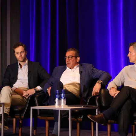 Three men in suits and business casual attire seated on stage chairs during a panel discussion with blue curtains behind.