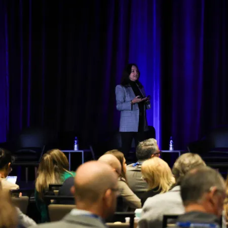 Speaker presenting on stage to an engaged audience in a dimly lit conference room with blue curtains.