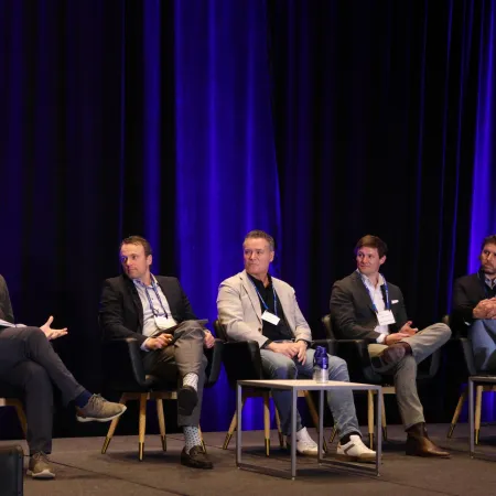 Five male panelists sitting on stage in front of blue curtains during a conference discussion.