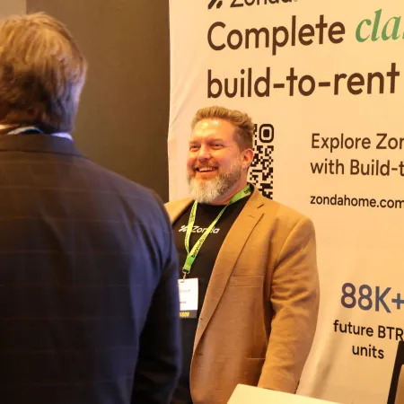 Smiling man in brown blazer talks with attendee at Zonda Build-to-Rent booth displaying promotional banner.