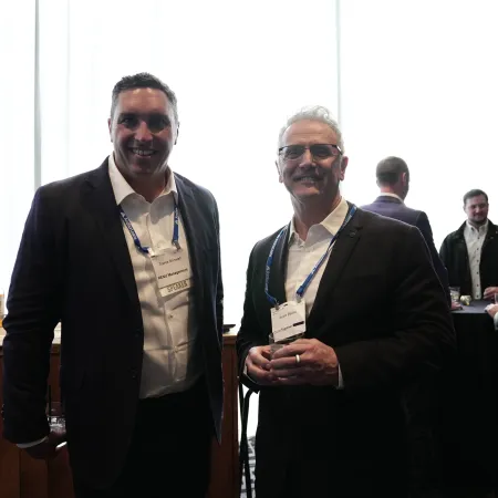Two men in business attire with name badges at a networking event in a well-lit room with other attendees.