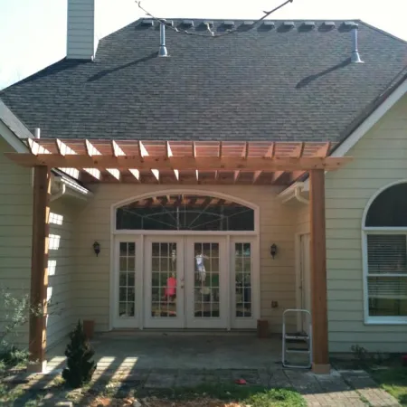 Wooden pergola attached to beige house with arched glass doors and a shingled roof under bright sunlight.