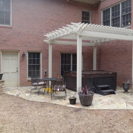 Backyard patio with pergola, hot tub, metal table and chairs, stone floor, and potted plants beside a brick house.