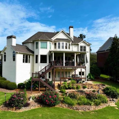Large white two-story house with wooden deck, outdoor stairs, and landscaped garden under blue sky.