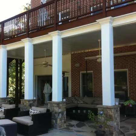 Outdoor patio area with stone pillars, white columns, ceiling fans, and cushioned seating under a wooden deck.