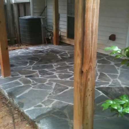 Covered outdoor patio with irregular stone flooring and wooden support posts beside a house wall.