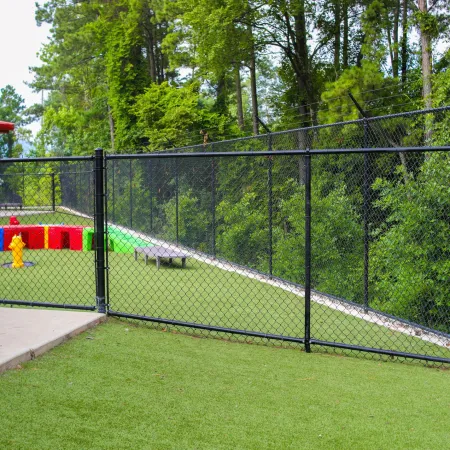 Black chain-link fence encloses a play area with artificial grass, colorful toys, and surrounding trees.