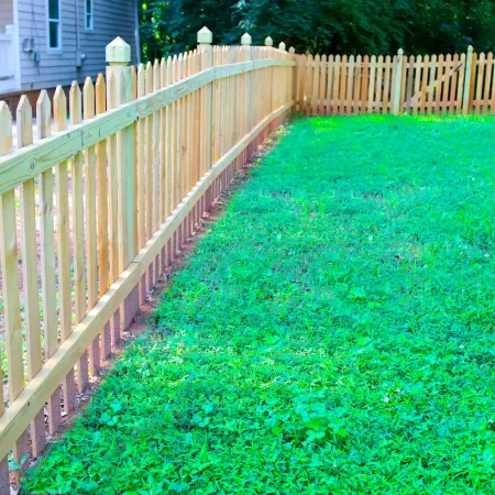 New wooden picket fence enclosing a green backyard next to a white house and porch.