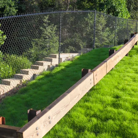 Green grassy slope bordered by wooden retaining wall and chain-link fence with stone steps behind it.