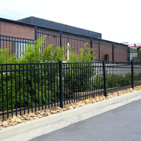 Black metal fence with vertical bars lining a driveway alongside green bushes and a brick building.
