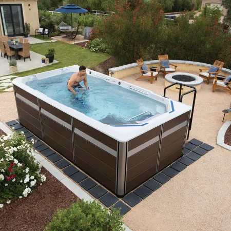 Man exercising in a backyard swim spa surrounded by garden, patio furniture, and flower beds on a sunny day.