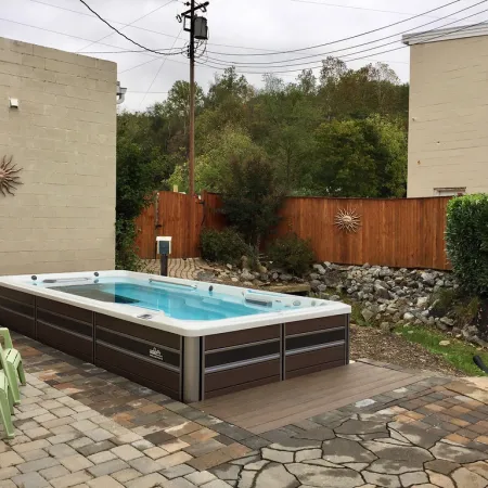 Outdoor patio with a rectangular swim spa, green chairs, and sun decorations on beige walls and wooden fence