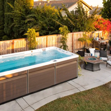 Backyard patio with a modern swim spa, outdoor seating, fire pit, and lush green plants under warm sunlight.